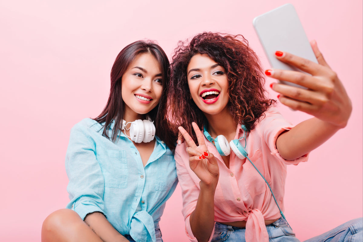 Two smiling women taking a selfie together with a smartphone against a pink background.