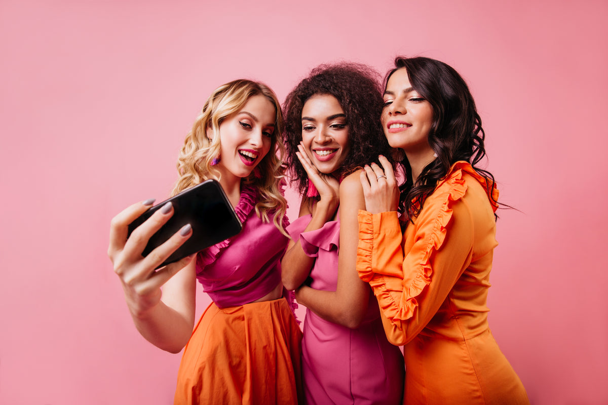 Three smiling women in colorful pink and orange outfits posing together for a selfie against a pink background.