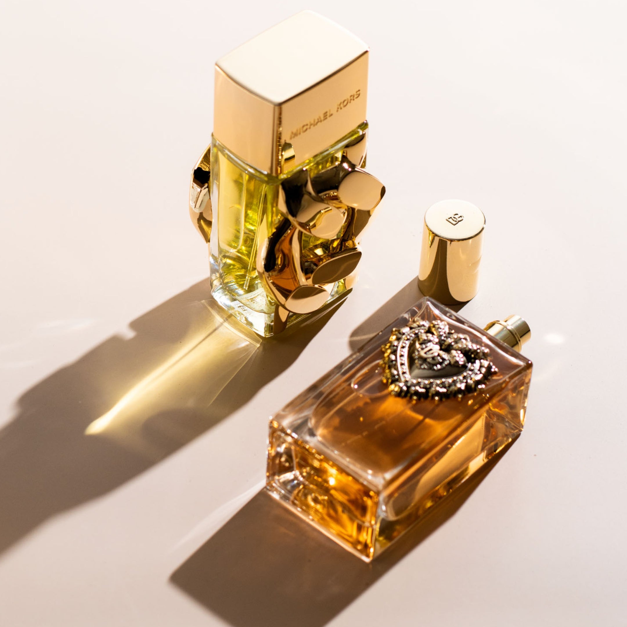 Michael Kors perfume bottle beside a Dolce & Gabbana fragrance with gold details on a light background.

