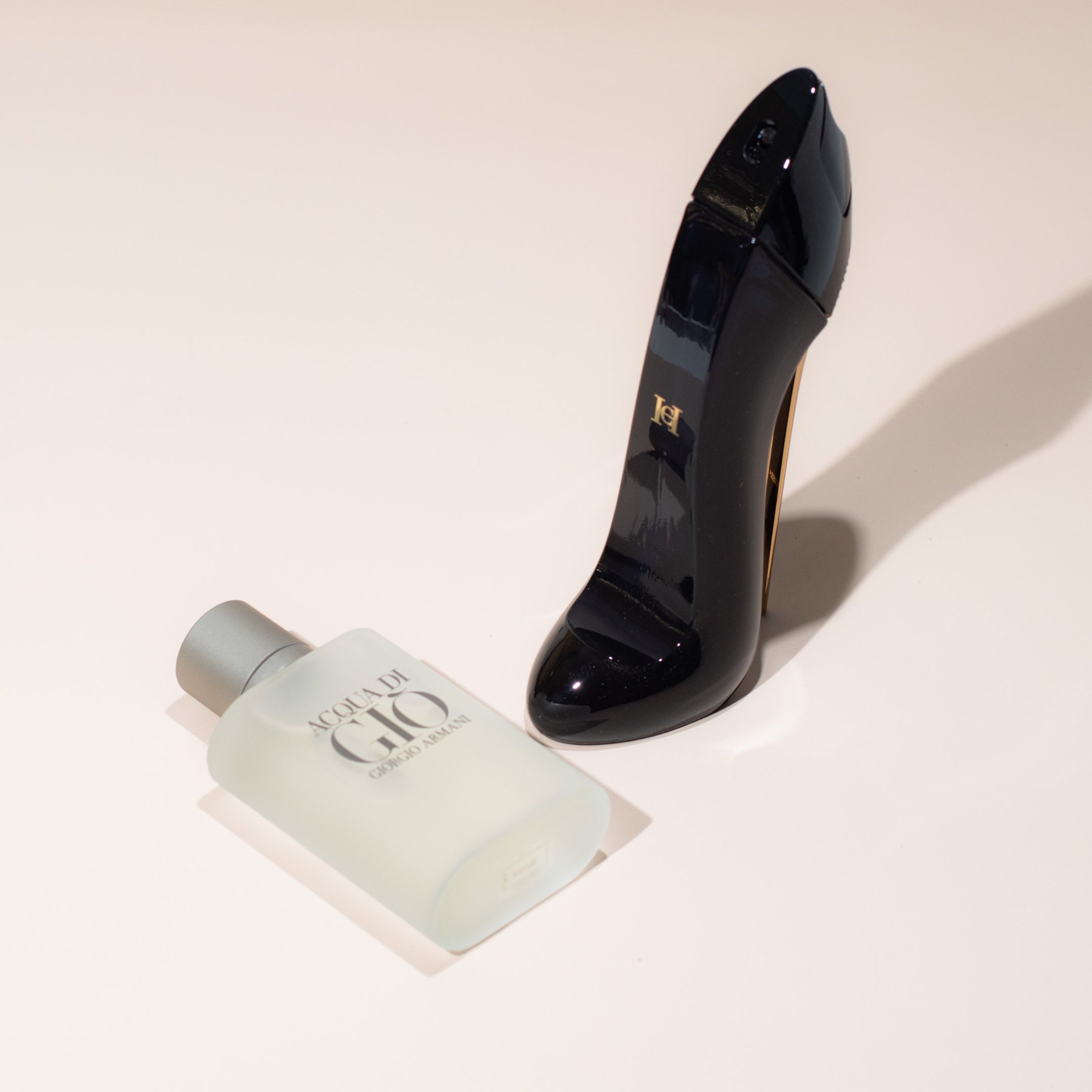 Giorgio Armani Acqua di Giò bottle next to Carolina Herrera Good Girl stiletto-shaped perfume on a light background.
