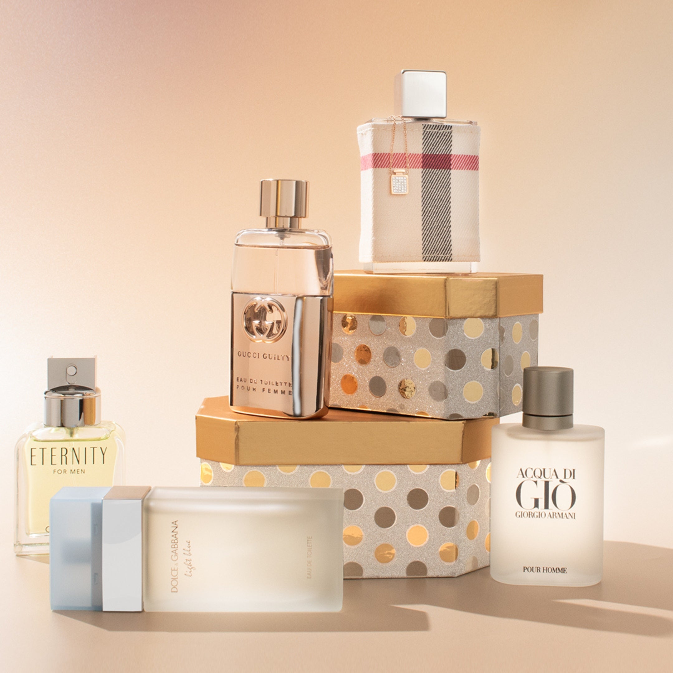 Assorted designer perfume bottles displayed on stacked gift boxes, featuring Gucci Guilty, Acqua di Giò, Burberry, Eternity, and Dolce & Gabbana fragrances.