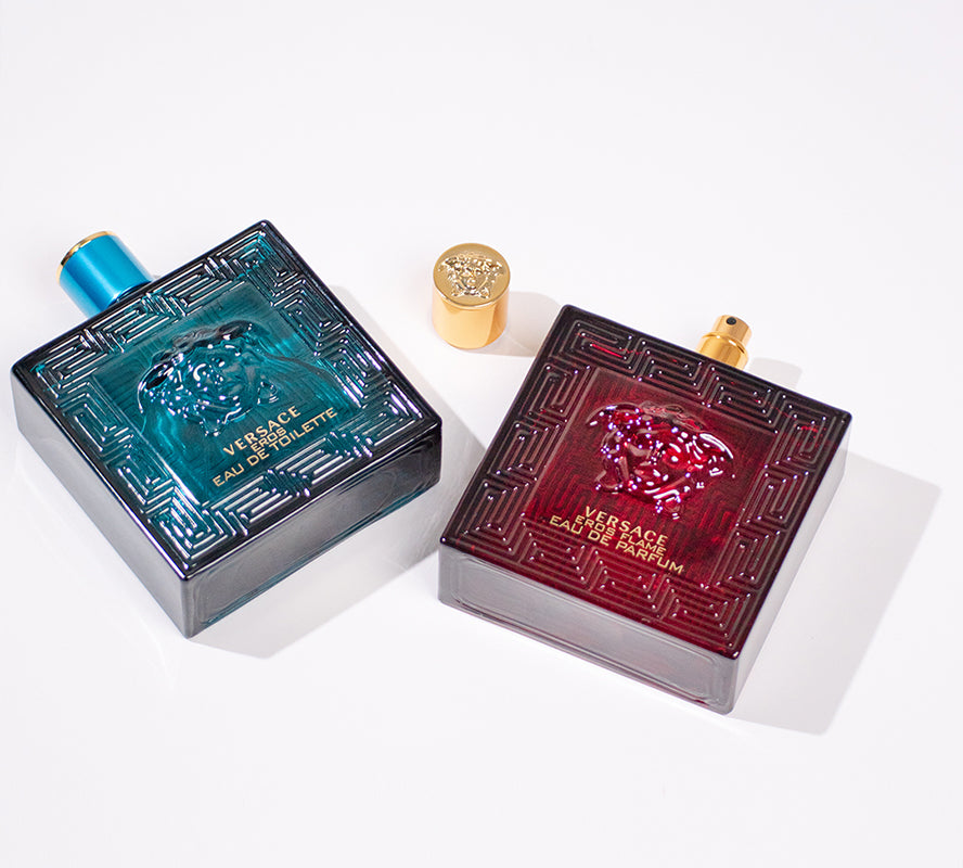 Two square Versace perfume bottles on a white surface: a teal “Eros” bottle on the left and a red “Eros Flame” bottle on the right, with a gold cap placed between them.