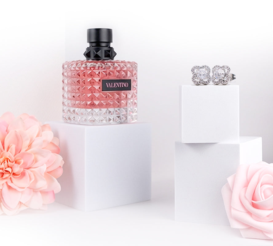 Pink Valentino perfume bottle and diamond earrings displayed on white blocks with soft pink flowers.